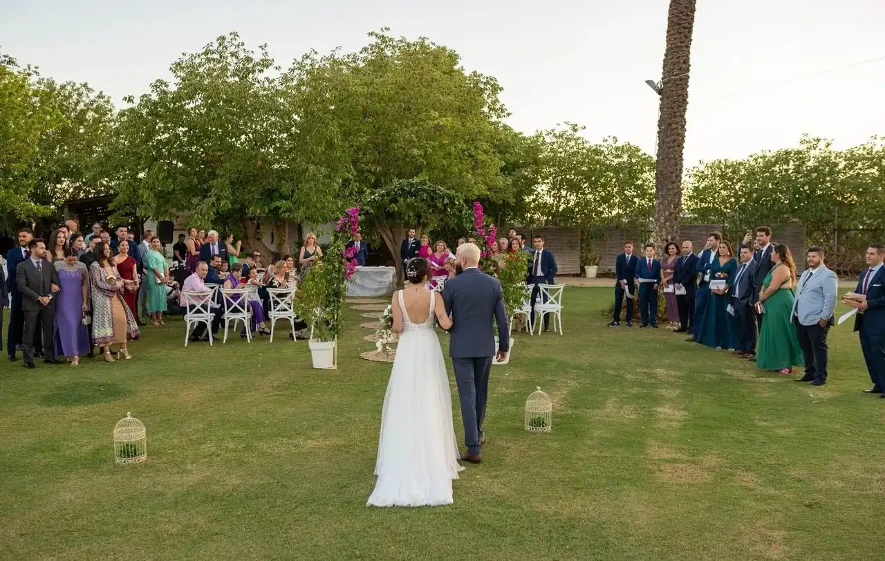 La novia del brazo de su padre camina sobre el césped hacia los invitados, con árboles al fondo
