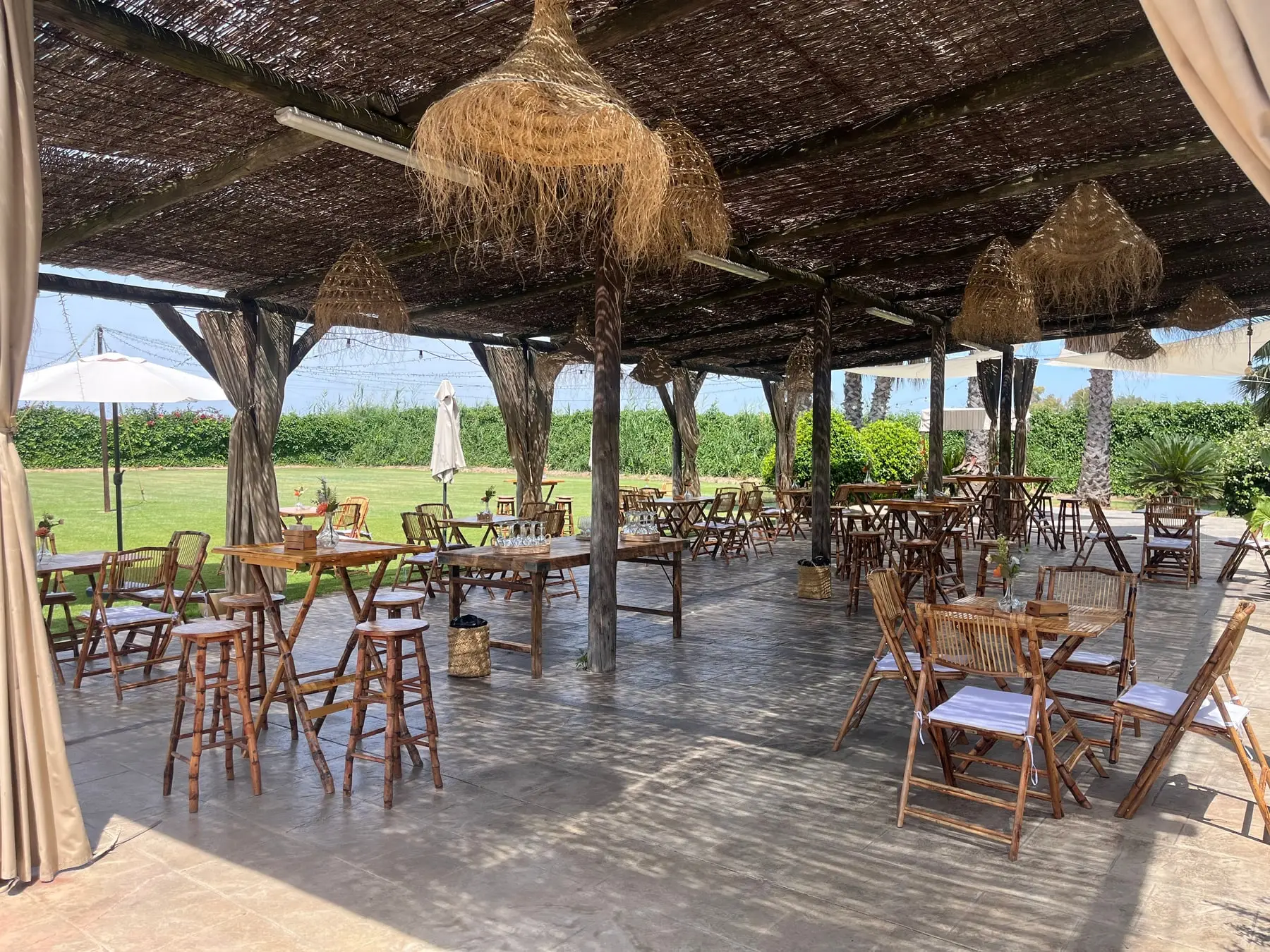 La terraza bajo el porche de esparto, con mesas altas, sillas de madera y lámparas de fibra, abierta al césped