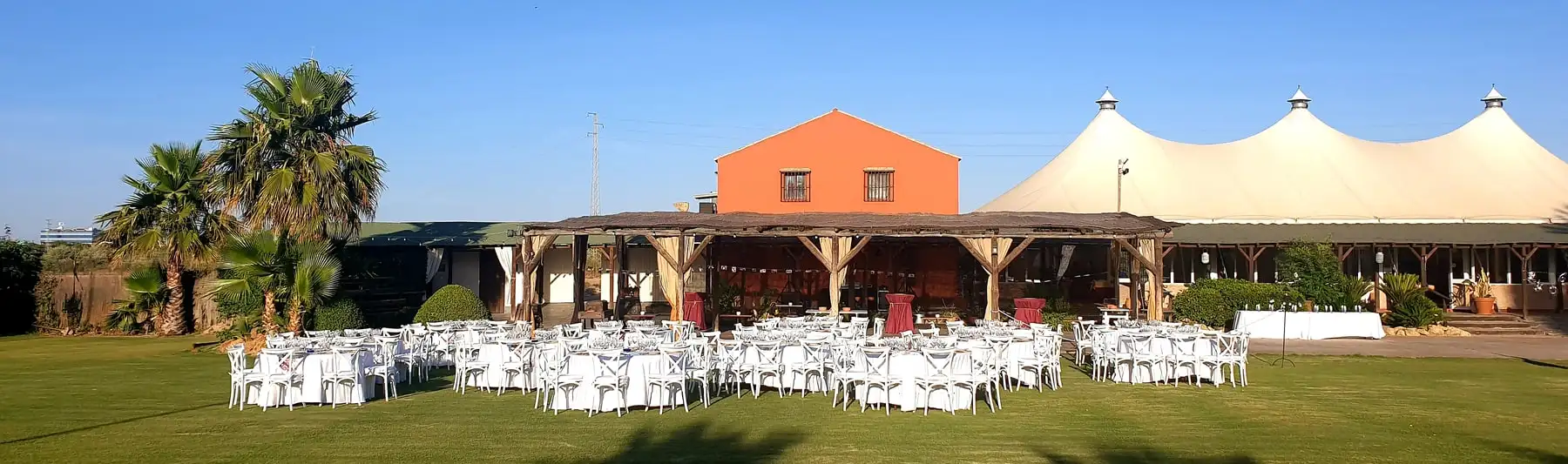 La pradera con las mesas redondas puestas, el porche de madera, la casa de color teja, las palmeras y la carpa blanca al fondo