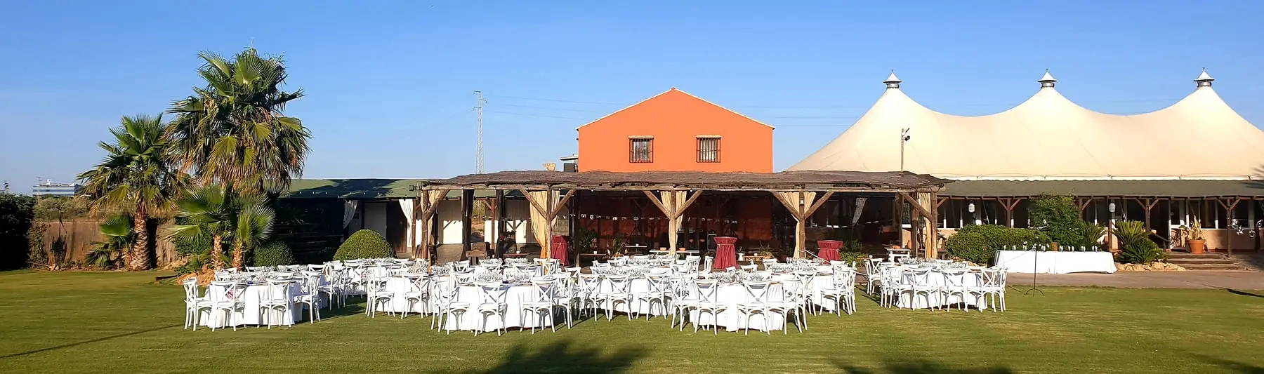 La pradera con las mesas redondas puestas, el porche de madera, la casa de color teja, las palmeras y la carpa blanca al fondo